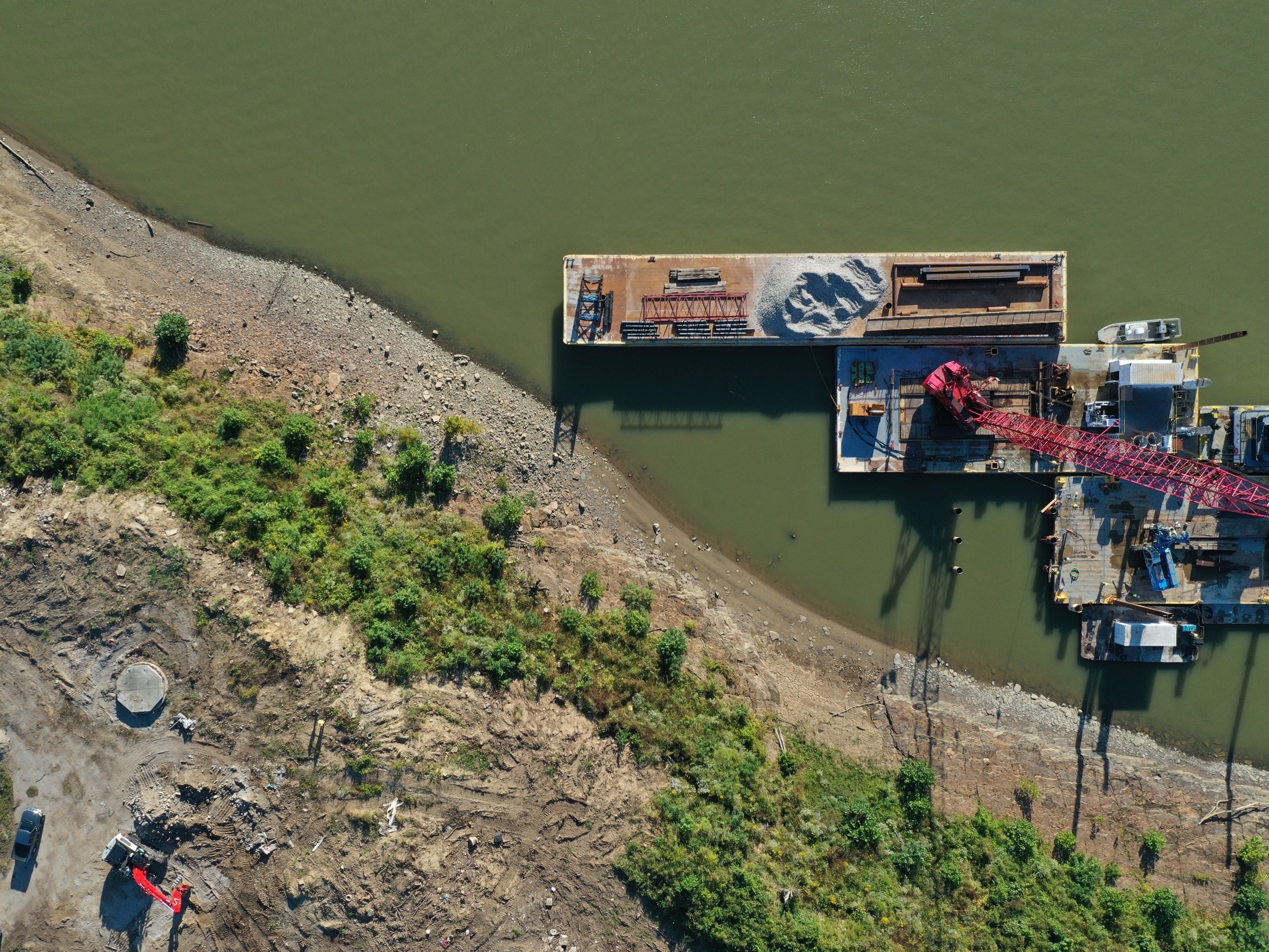 Aerial view of a barge on a greenish river with a red crane, gravel piles, and a rocky, vegetated shoreline nearby.