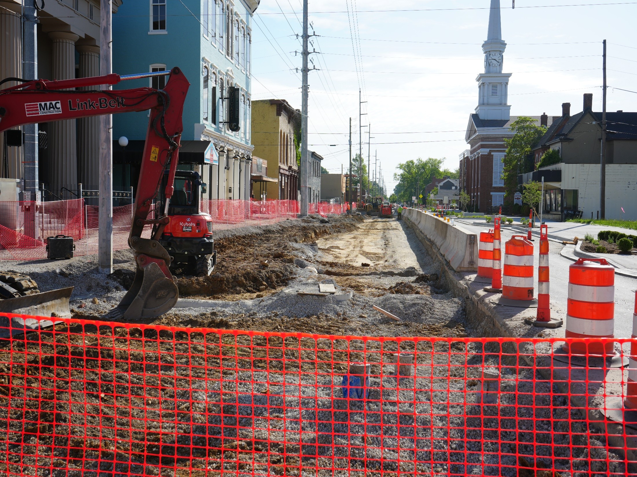 Construction site along a city street with orange safety barriers, a red excavator, a muddy trench, and a distant white church steeple.