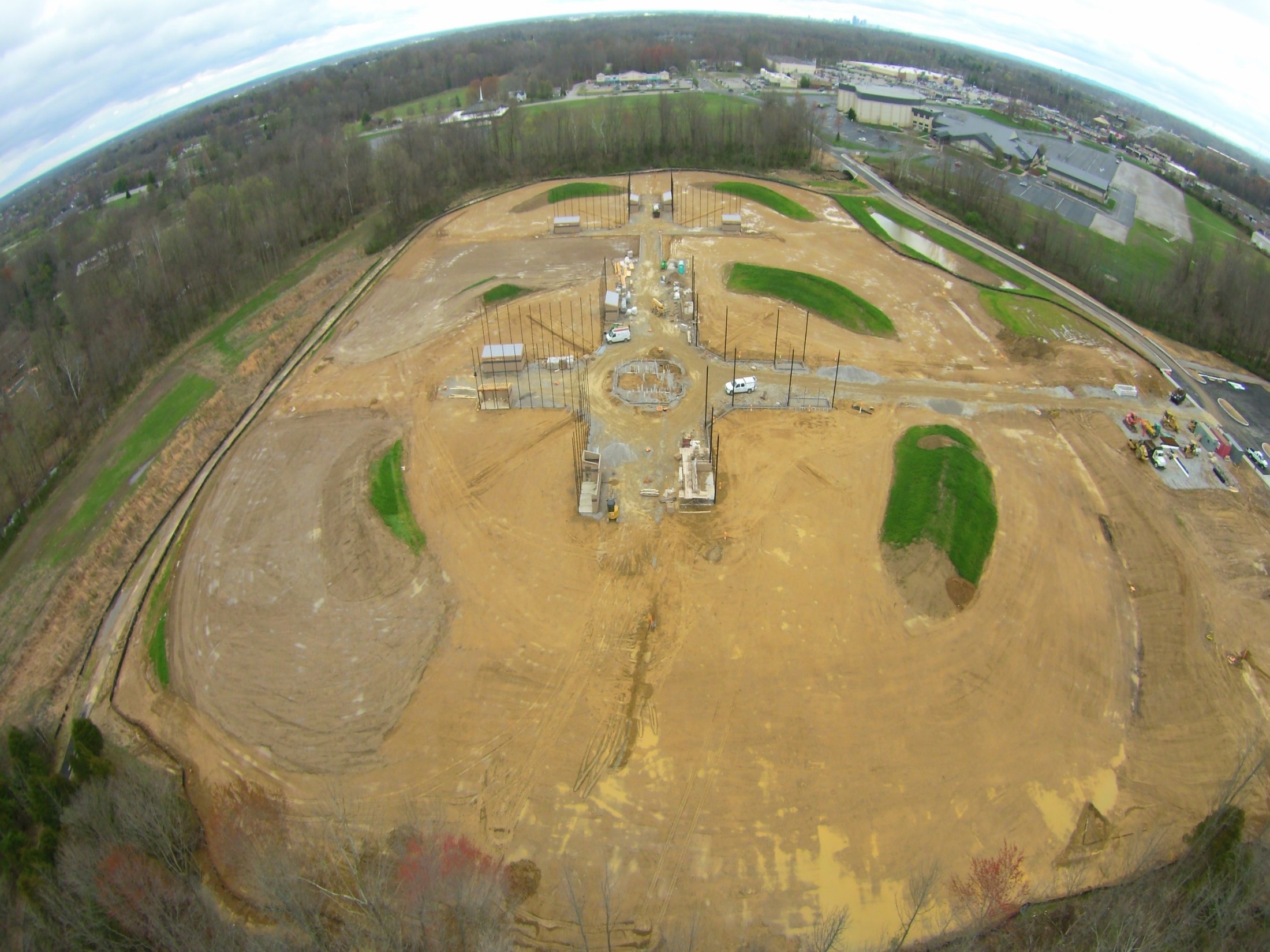 Aerial view of a construction site with circular dirt areas, scaffolding, and exposed foundations surrounded by trees and a road network.