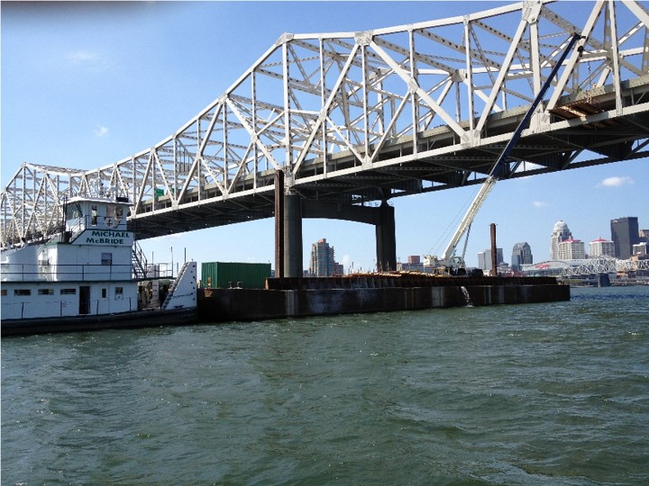 White steel truss bridge spans a river, with a docked boat labeled 'Michael McBride' and a distant city skyline.