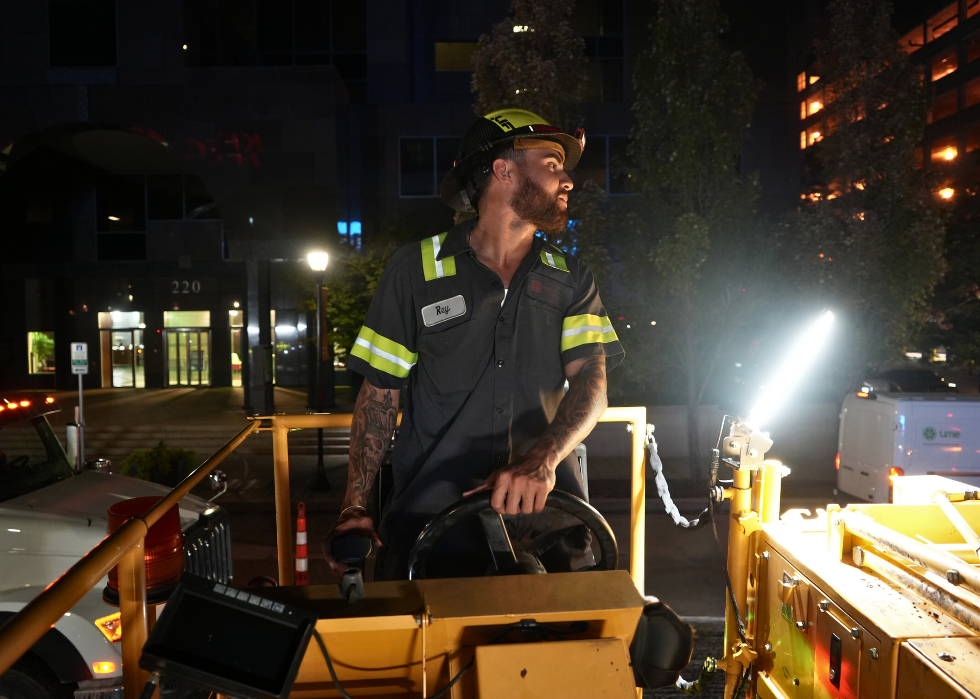 Bearded male construction worker in a yellow helmet and reflective safety gear at night, standing beside a yellow utility vehicle.