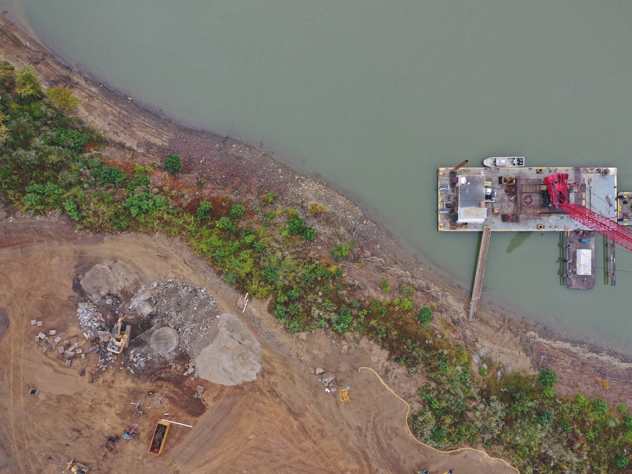 Aerial view of a construction site along a muddy shoreline with a docked barge and red crane extending from the platform into the water
