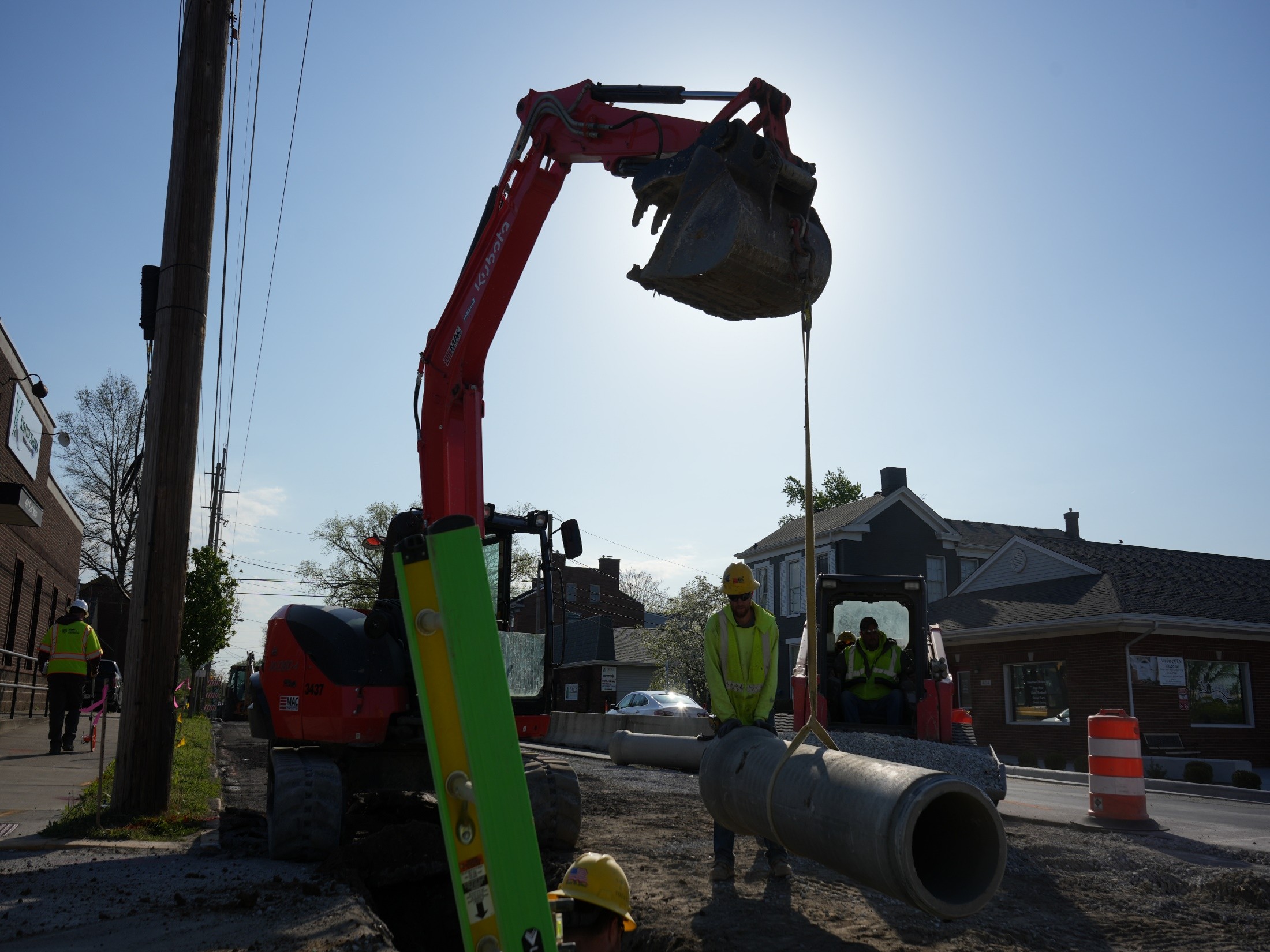 Red excavator bucket lifts a large gray pipe across a street, with workers in safety vests nearby and the sun behind the equipment.