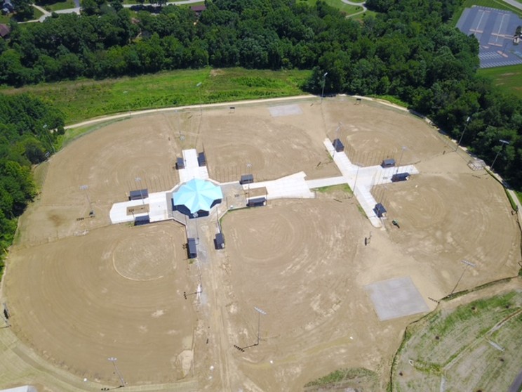Aerial view of a circular dirt field with a blue-roof pavilion and white walkways at a park center, surrounded by trees.