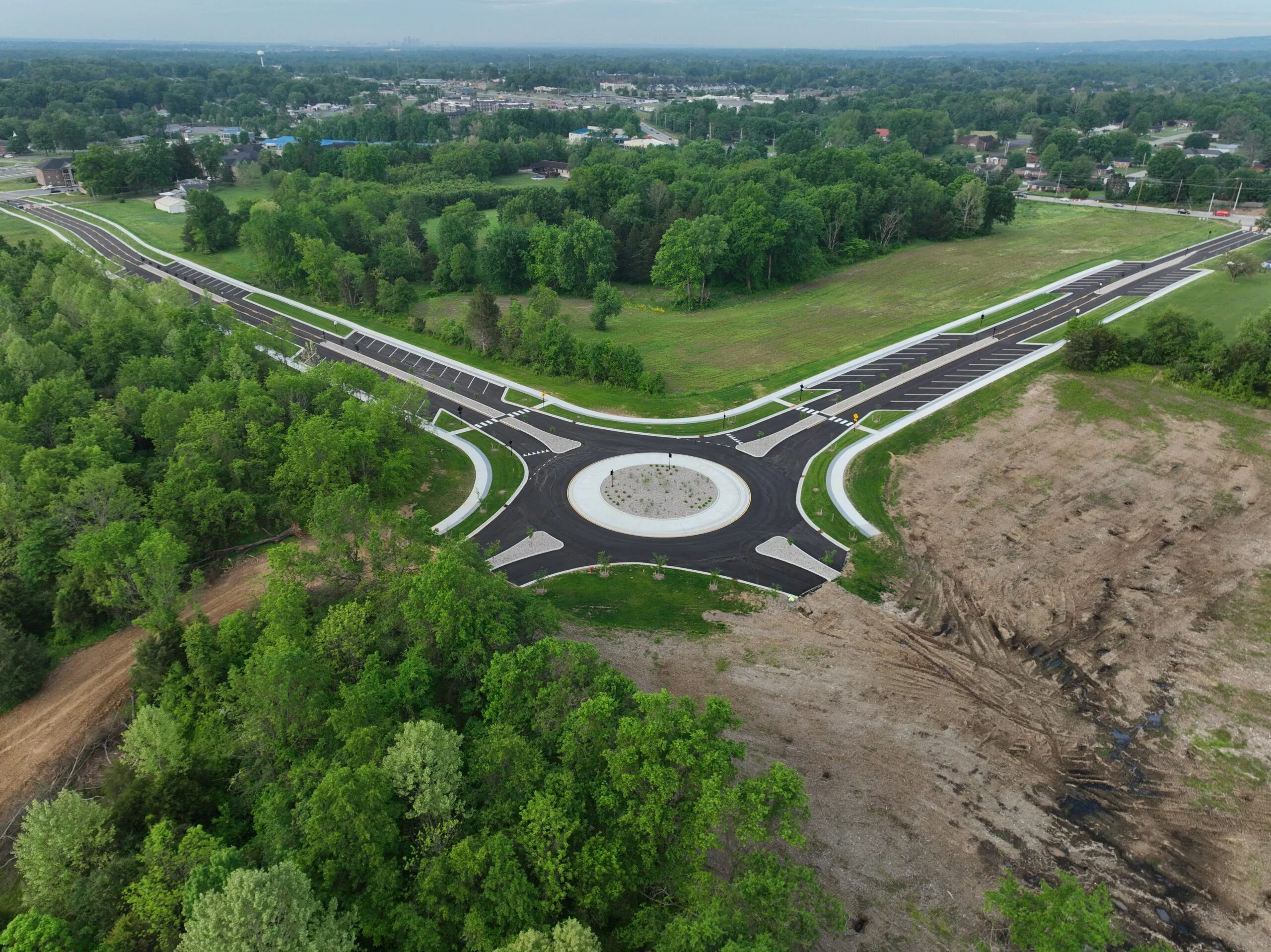 Aerial view of a large roundabout interchange under construction, with multiple curving roads, white curbs, and a circular central island surrounded by trees and green space.