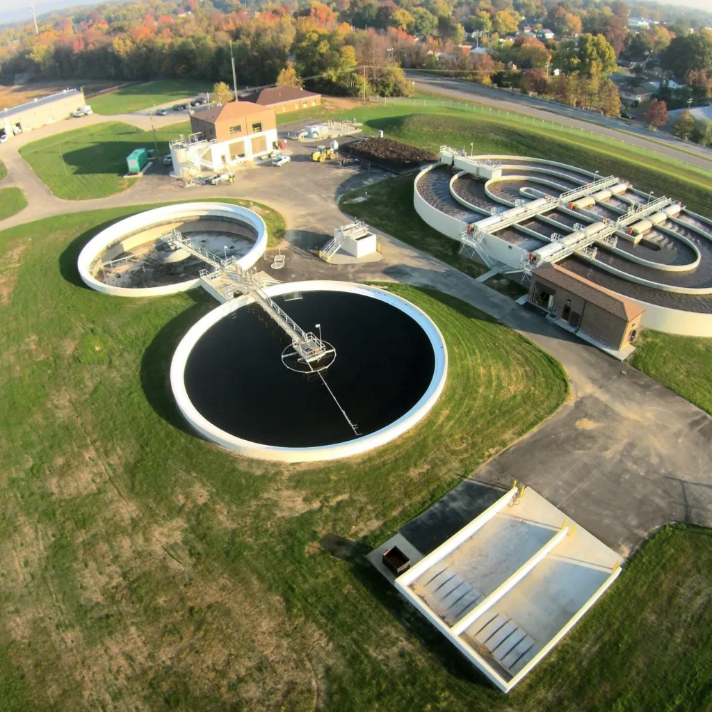 Aerial view of a wastewater treatment facility with circular sedimentation tanks and interconnected basins amid green fields and roads nearby.