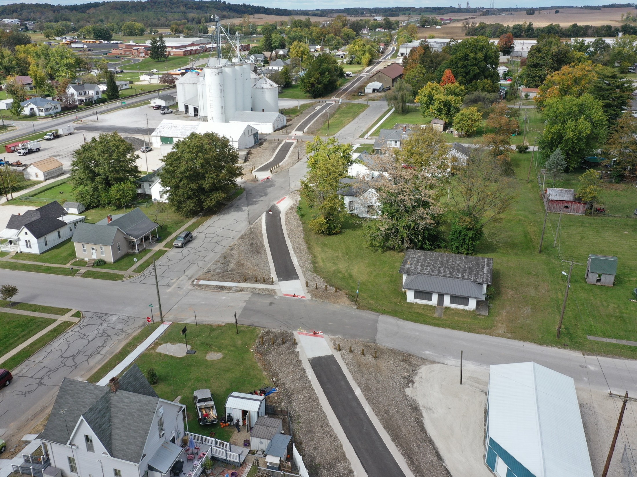Aerial view of a small town with a central road, white silo complex, and surrounding houses and trees.