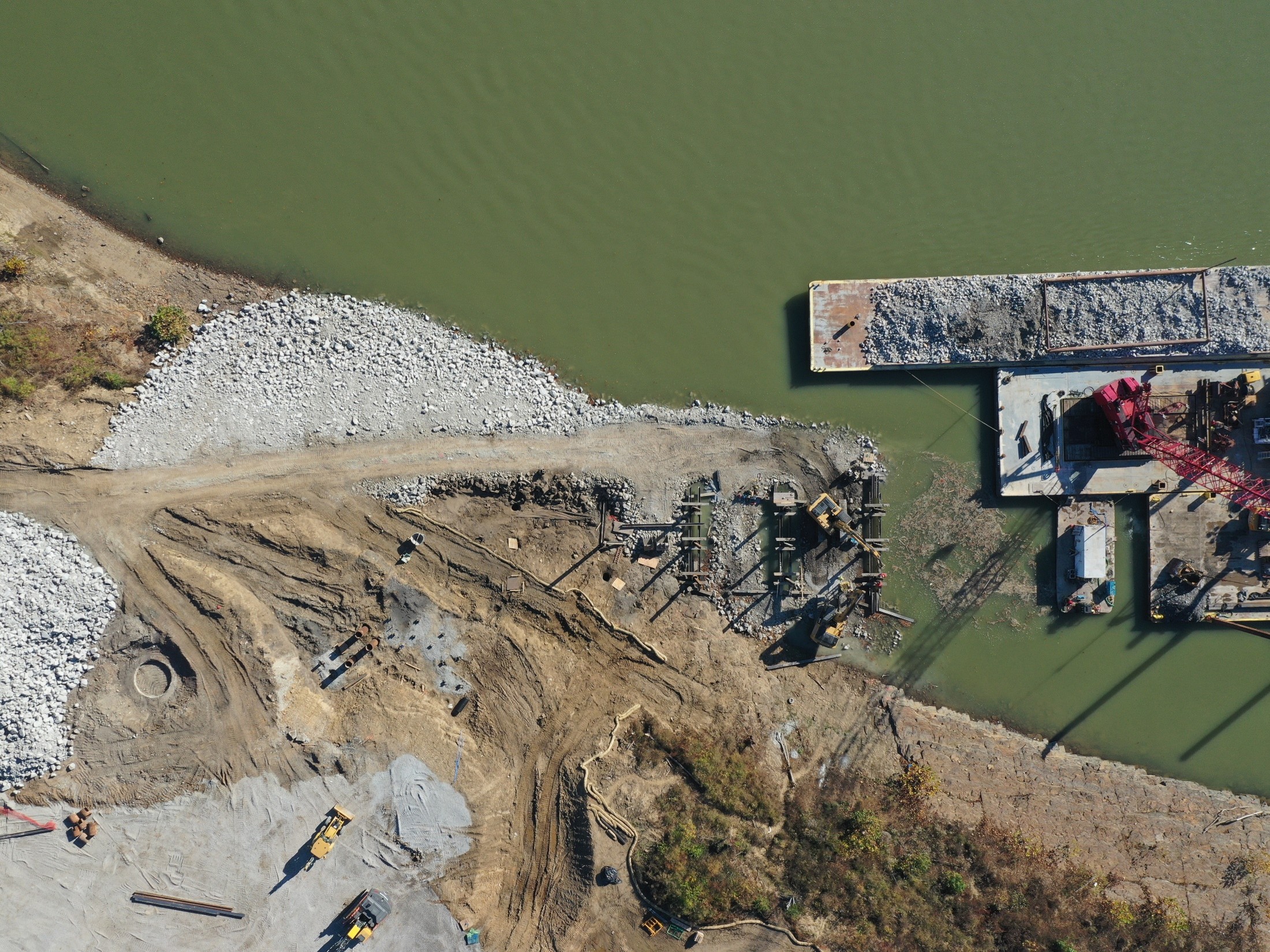Aerial view of a construction site along a green river, with a barge loaded with gravel docked at a concrete platform.