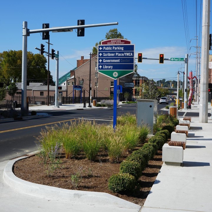 Blue downtown directional sign listing Public Parking, Scribner Place/YMCA, Government Center, and Library, with a sidewalk and traffic lights in a sunny street scene.