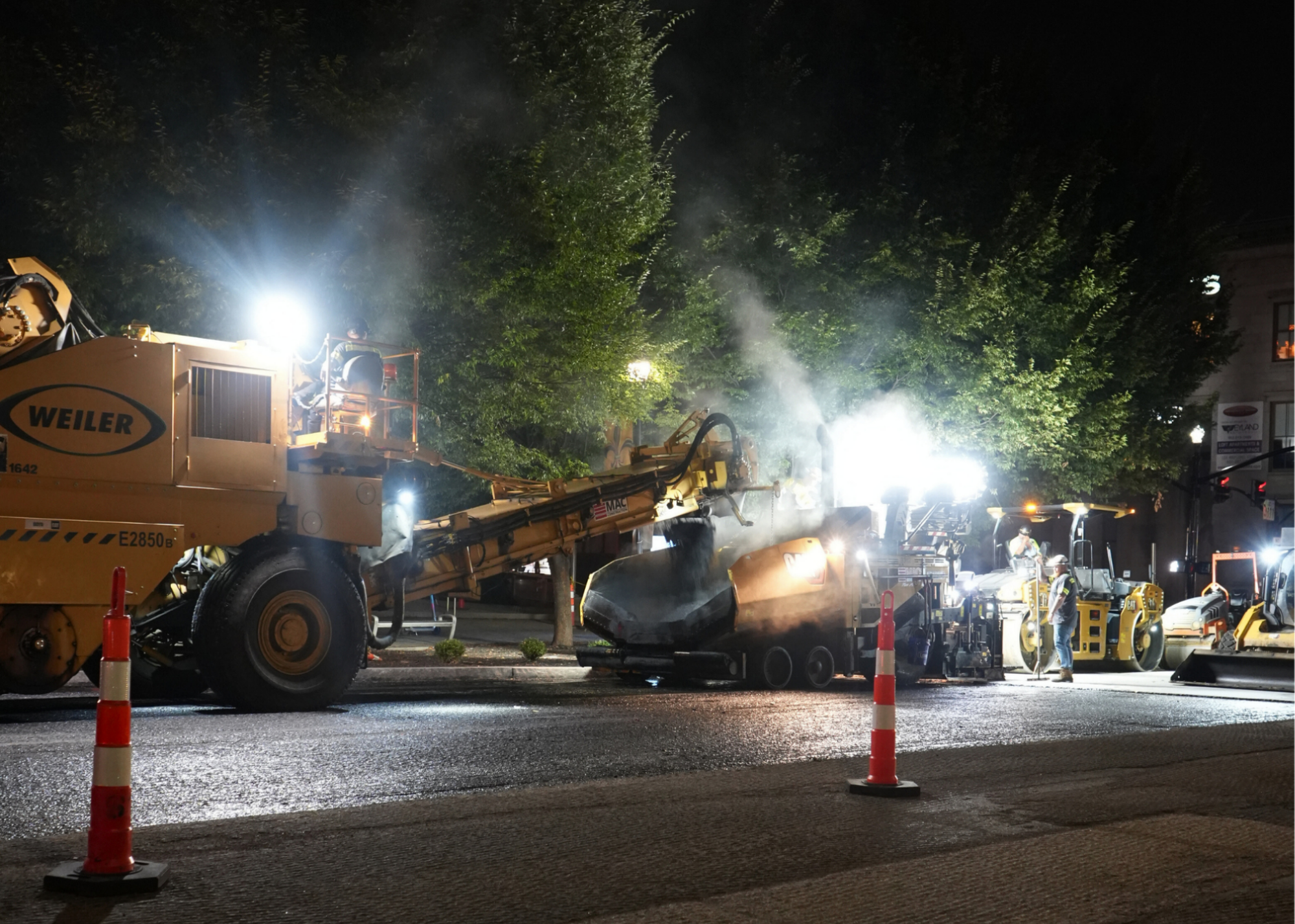 Nighttime construction scene with a large yellow crane truck labeled WEILER, workers on the platform, and orange safety cones along the street.