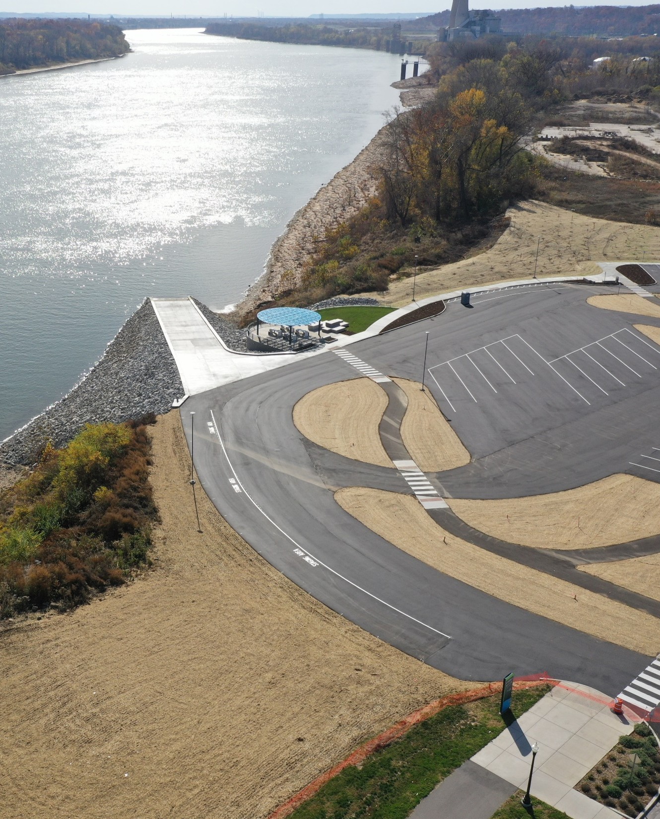 Aerial view of a riverside park with a curved road, parking lot, and a blue-canopied picnic area by the water.