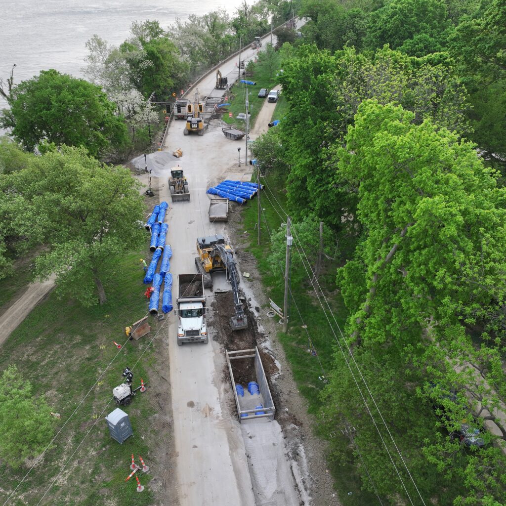 Aerial view of a riverfront road construction site with heavy machinery, dump trucks, orange cones, and blue tarps along a tree-lined bank.