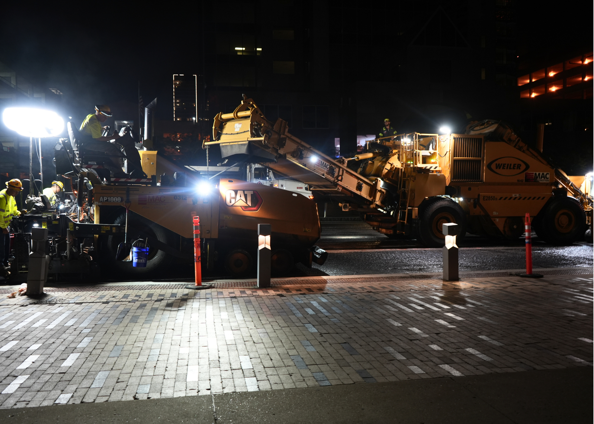 Nighttime construction site with a large yellow excavator loading debris onto a truck, workers in reflective gear illuminated by bright floodlights.