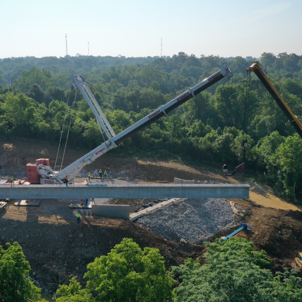 Crane sections extend over a rocky excavation site with trees in the background and workers nearby.