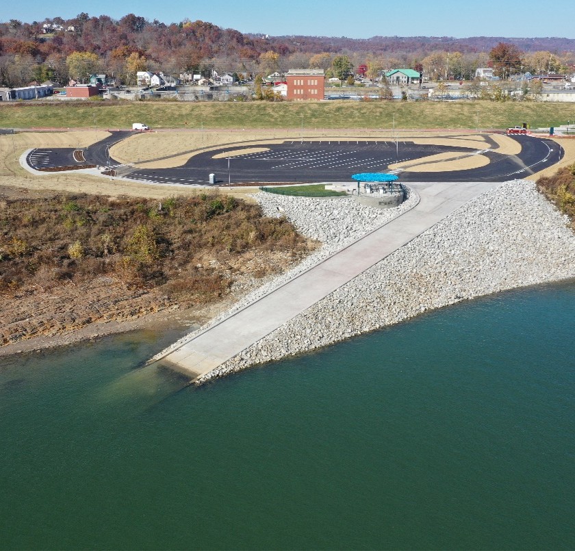 Aerial view of a circular driving track with a surrounding grassy area, parked cars, and a small town in the distance by a river.