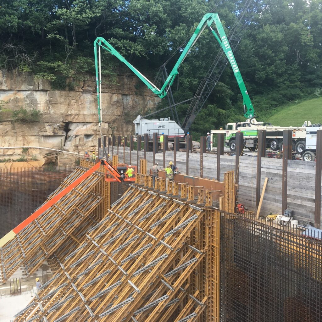 Construction site showing a large green concrete pump arm over wooden rebar formwork with workers in hard hats nearby.