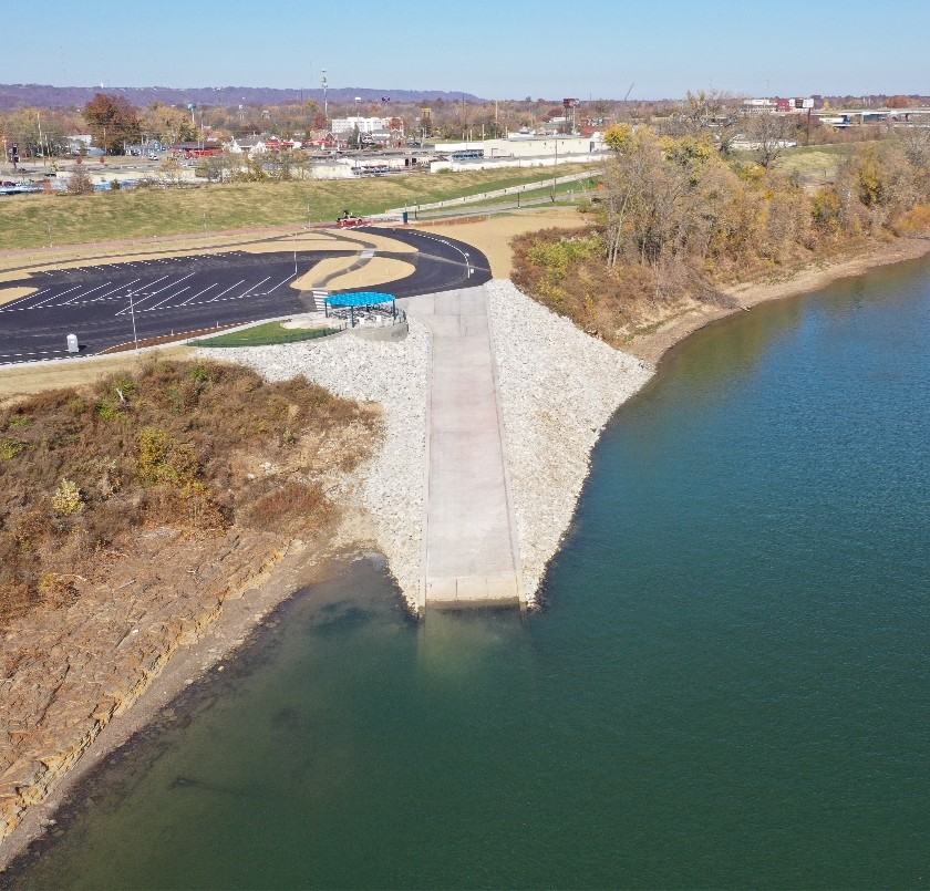 Aerial view of a riverbank with a concrete pier extending into the water, rocky shoreline, and a paved walkway; a curved parking lot and small blue shelter are visible in the background amid fall-colored trees and a town skyline far off.