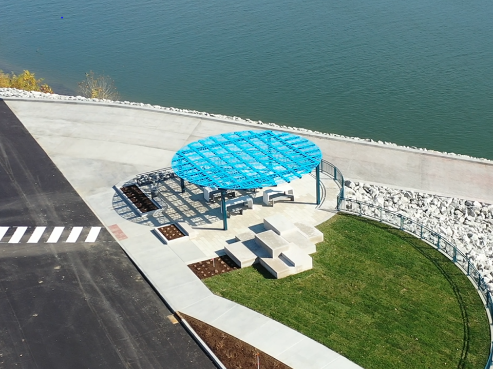 Aerial view of a waterfront seating area with a blue circular lattice canopy over concrete tables and benches beside a grassy circle and stone barrier.