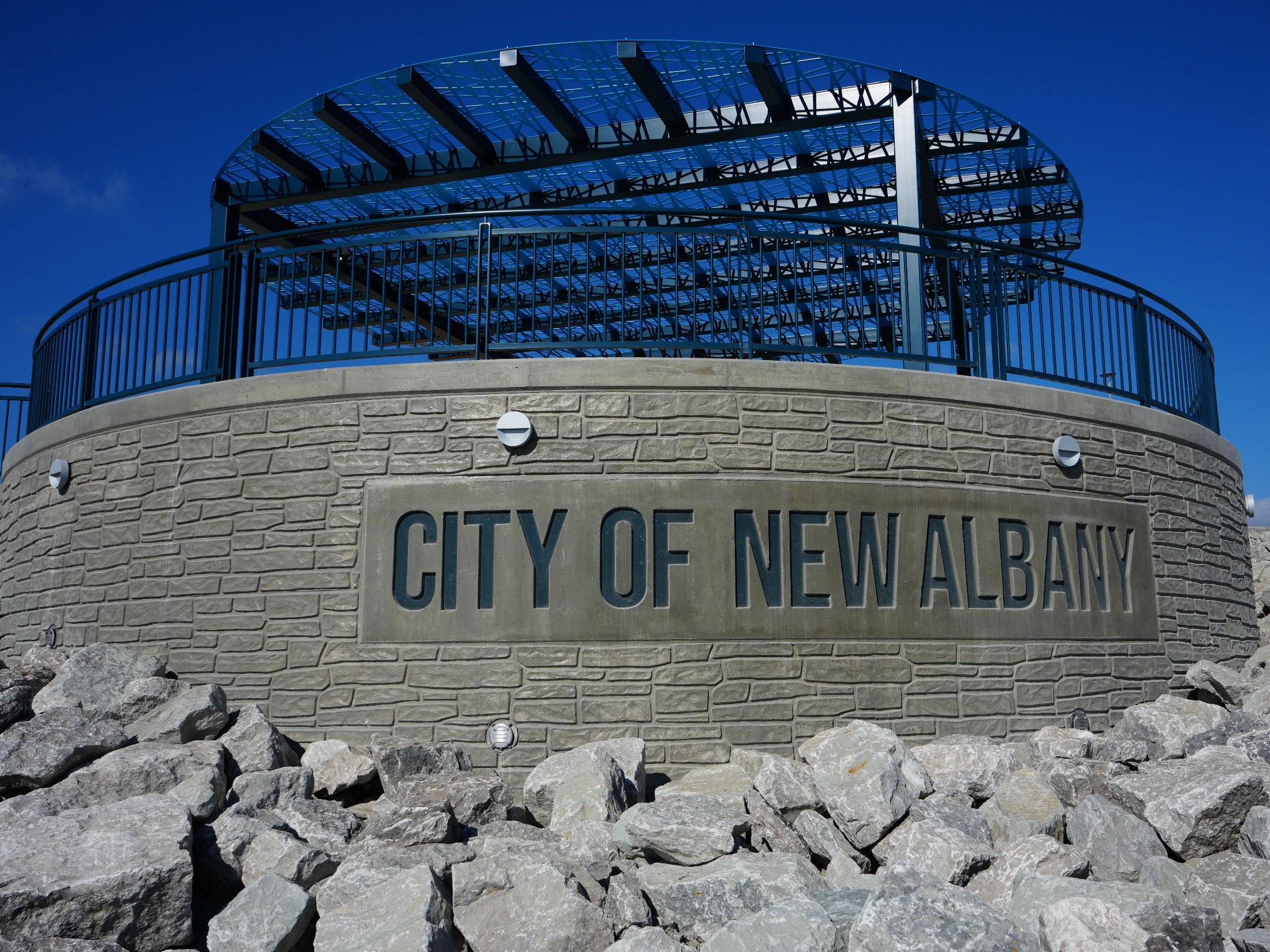 Curved stone wall with a blue railing surrounding a circular metal framework structure against a clear blue sky; sign reads 'CITY OF NEW ALBANY'