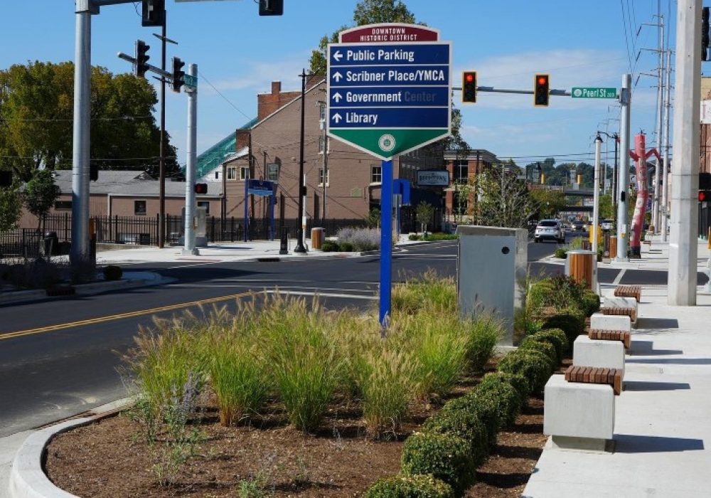 Blue downtown directional sign listing Public Parking, Scribner Place/YMCA, Government Center, and Library, with a sidewalk and traffic lights in a sunny street scene.